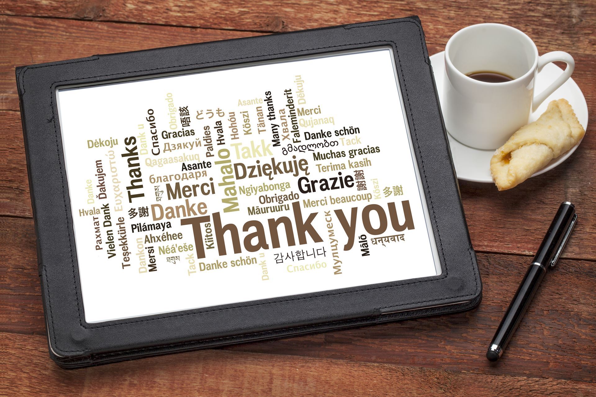 Thank you in many languages on a tablet with a cup of coffee, pastry, and a pen. 